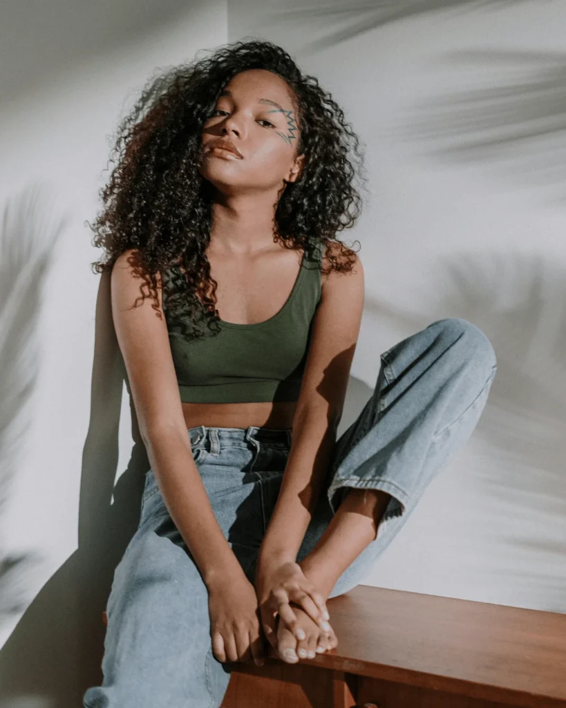 slim black woman with curly hair sitting on a wooden bench indoors. Shadows of plants behind her on a plain grey wall. 