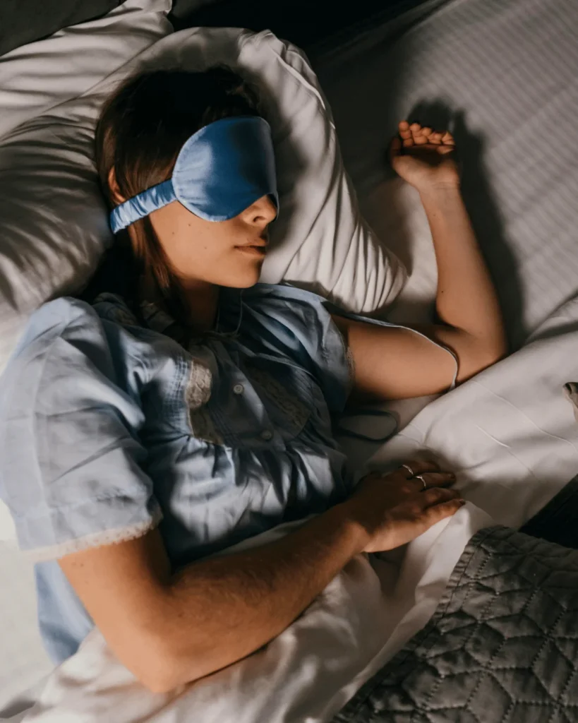 young white woman sleeping in a bed wearing blue satin eye mask. Lighting is dim. Pillow Method